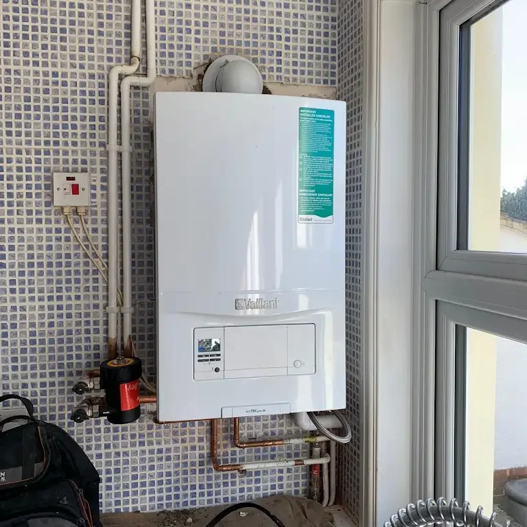 A white Vaillant boiler is mounted on a wall with blue and white mosaic tiles. It is connected to several pipes and valves. A window is to the right, allowing natural light into the space. An electricity switch is mounted nearby, and a black bag rests on the floor.