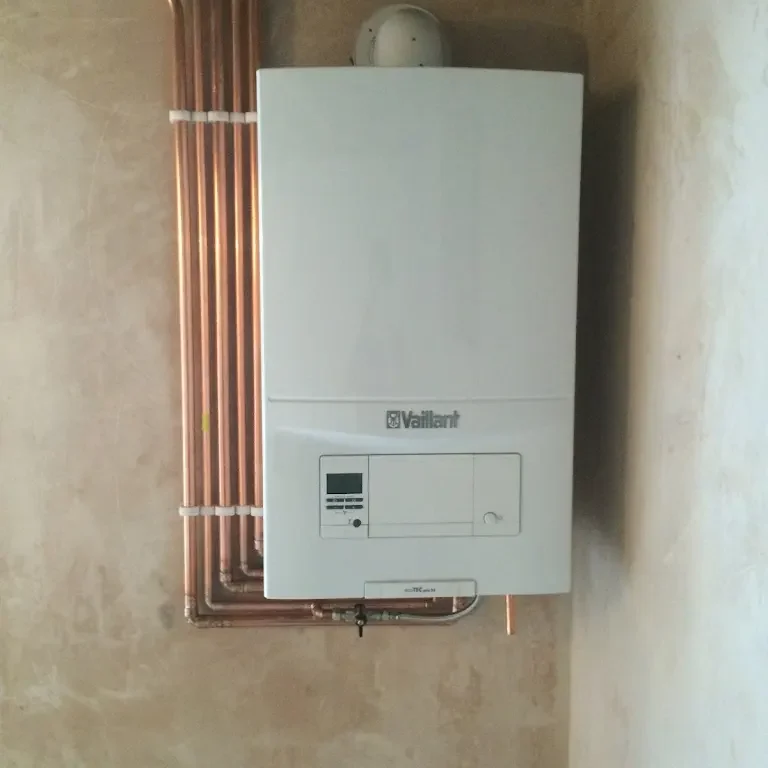 The image shows a wall-mounted Vaillant boiler installed with neatly arranged copper pipes along the wall. The boiler's control panel is visible, and it is installed on a plain, beige-colored wall.