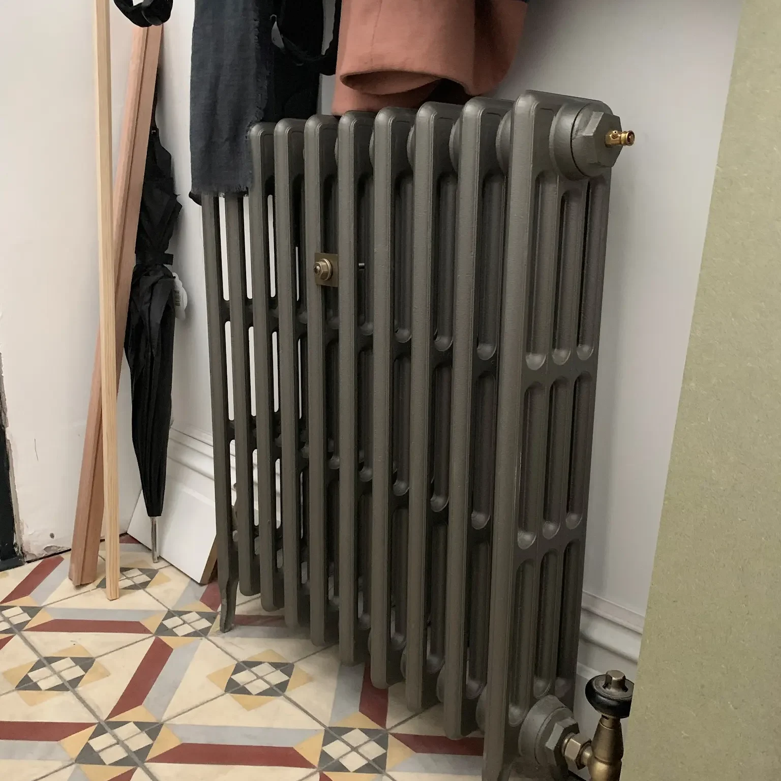 A vintage cast iron radiator is situated against a wall, with coats hanging above it. Pieces of wooden planks are leaning against the wall to the left. The floor features a patterned tile design in red, white, and yellow colors.