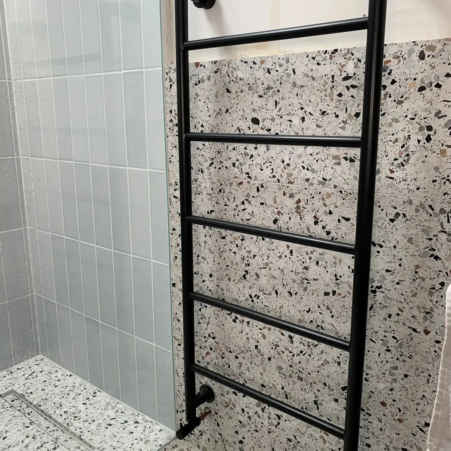 A bathroom corner featuring a sleek black towel rack with horizontal bars, mounted on a terrazzo wall. The adjacent section of the wall has white tiles with light gray grout. The shower area includes a white tiled shower base and glass enclosure.