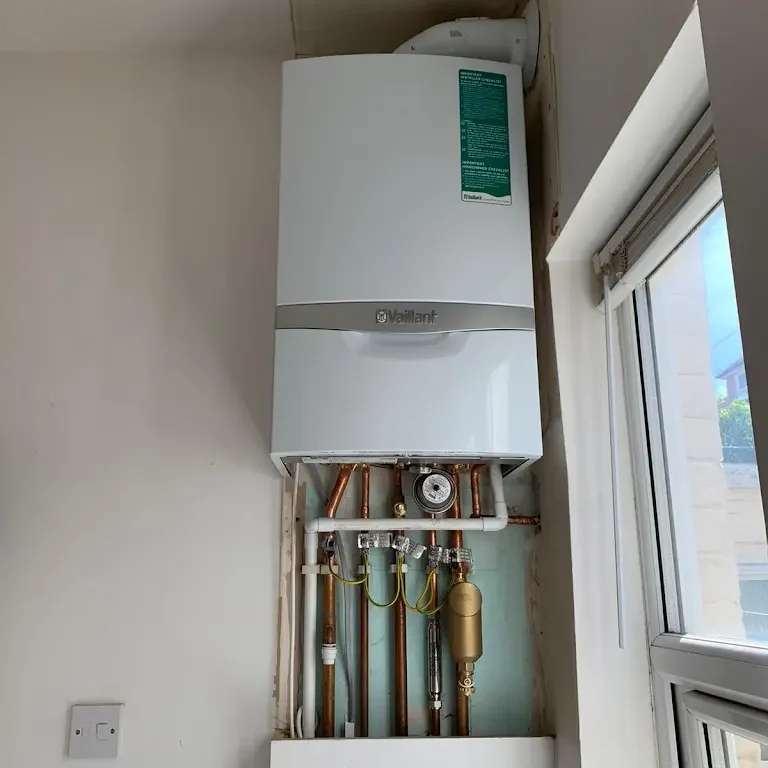 A wall-mounted gas boiler is installed in a bright room next to a window. It has visible pipes and connections beneath it, neatly arranged. The boiler appears clean and new, and the room has a white and clean appearance.